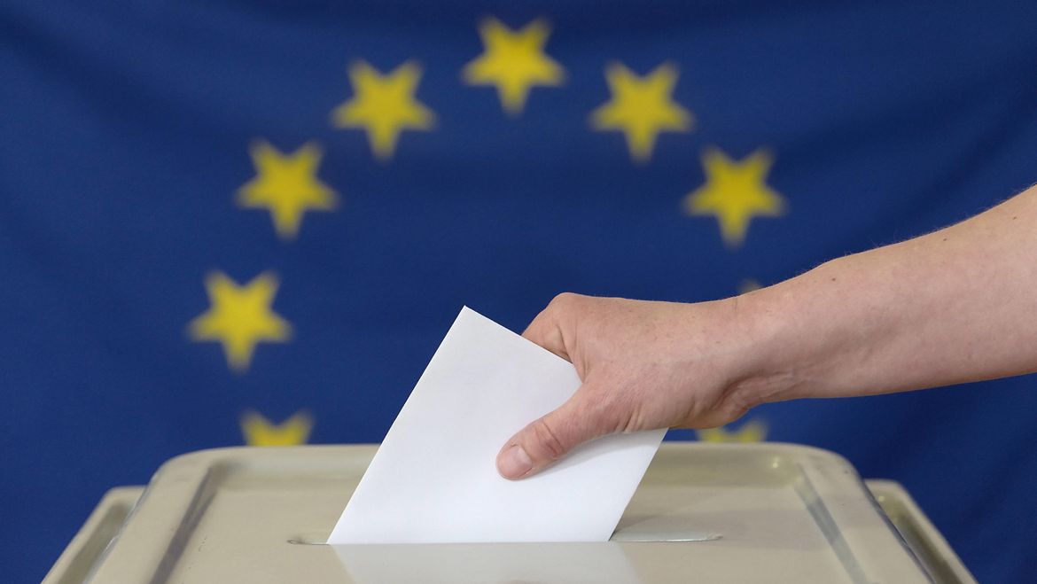 Eine Hand steckt einen Umschlag in eine Wahlurne vor der Europafahne.